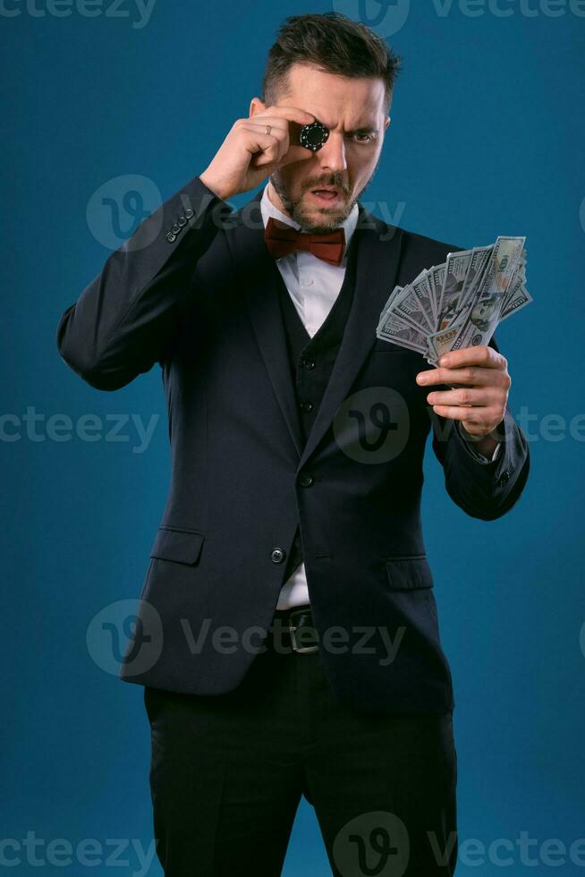 Man in black classic suit and red bow-tie showing one chip and some dollar bills, posing on blue background. Gambling, poker, casino. Close-up. photo