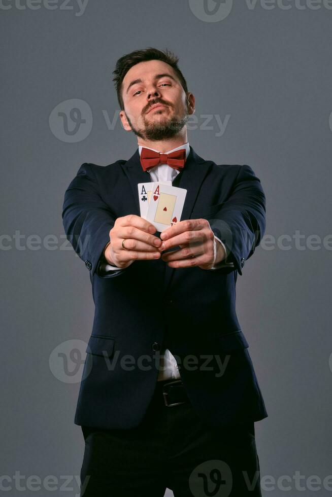 hombre en negro clásico traje y rojo corbata de moño demostración dos jugando tarjetas mientras posando en contra gris estudio antecedentes. juego, póker, casino. de cerca. foto