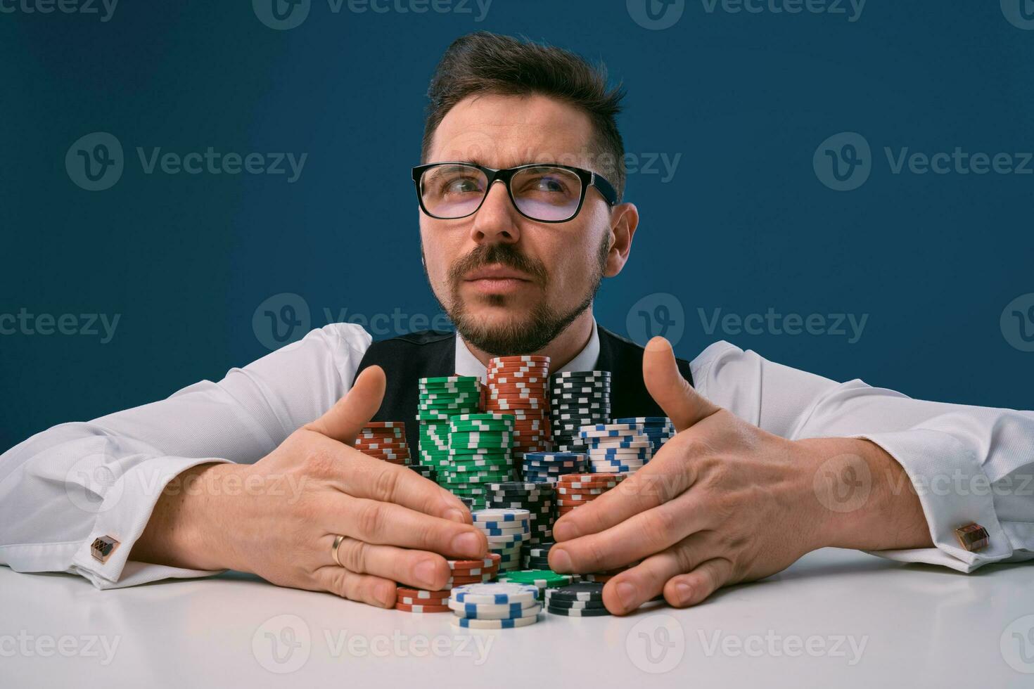 Guy in glasses, black vest and shirt sitting at white table with stacks of chips on it, posing on blue background. Gambling, poker, casino. Close-up. photo