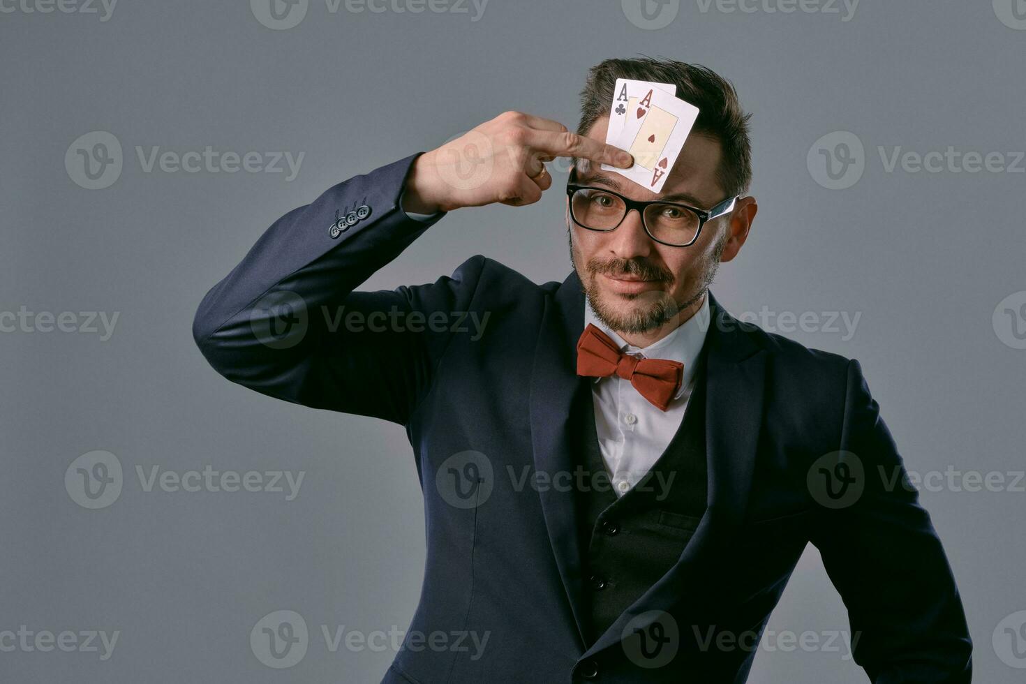 hombre en negro clásico traje, rojo corbata de moño, glases es demostración dos jugando tarjetas, posando en gris estudio antecedentes. juego, póker, casino. de cerca. foto