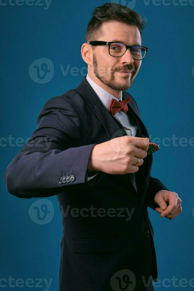 Man in black classic suit, red bow-tie, glases is showing two colored chips, posing on blue studio background. Gambling, poker, casino. Close-up. photo