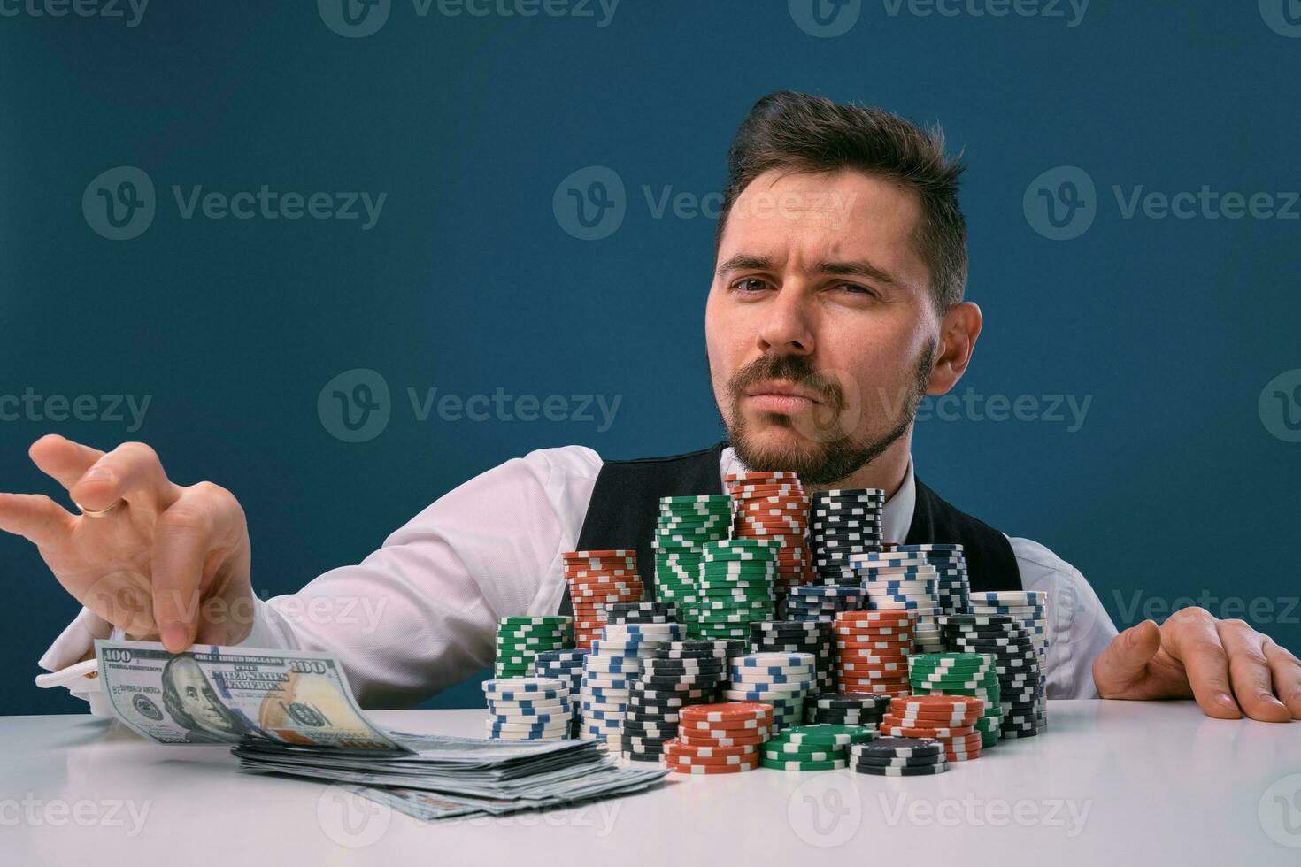 Man in black vest and shirt sitting at white table with stacks of chips and cash on it, posing on blue background. Gambling, poker, casino. Close-up. photo