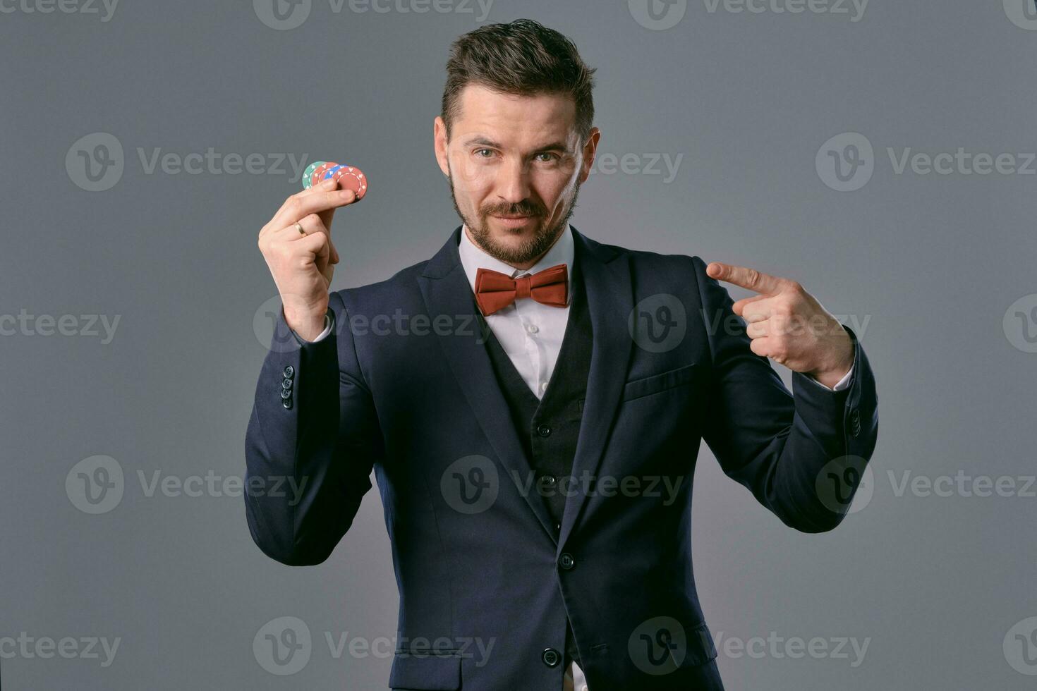 Man in black classic suit and red bow-tie showing some colored chips, posing against gray studio background. Gambling, poker, casino. Close-up. photo