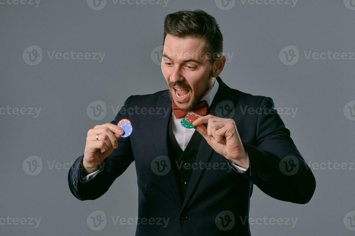 Man in black classic suit and red bow-tie showing some colored chips, posing against gray studio background. Gambling, poker, casino. Close-up. photo