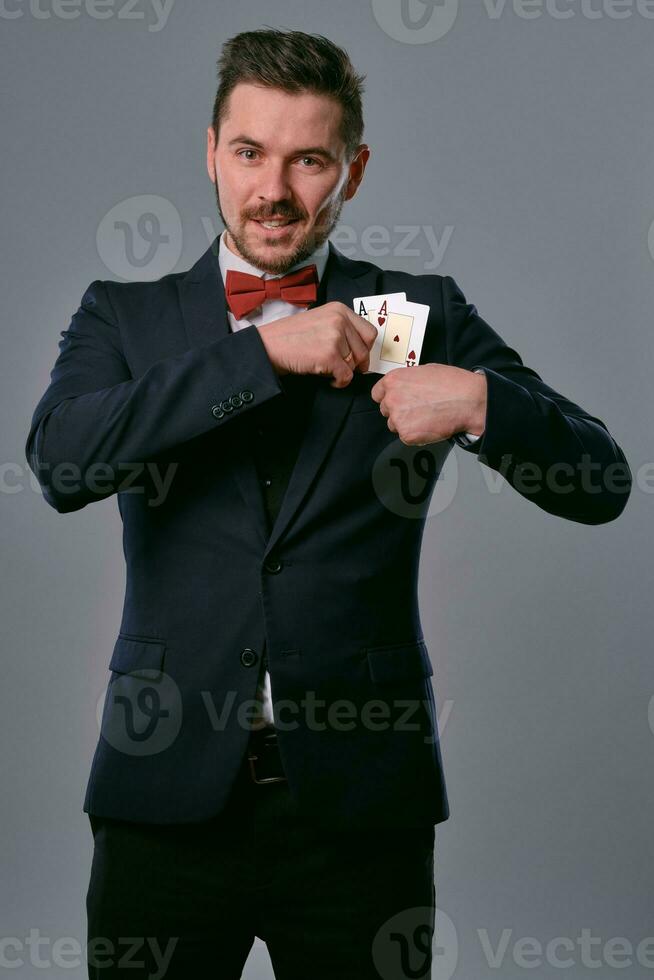 Man in black classic suit and red bow-tie showing two playing cards while posing against gray studio background. Gambling, poker, casino. Close-up. photo