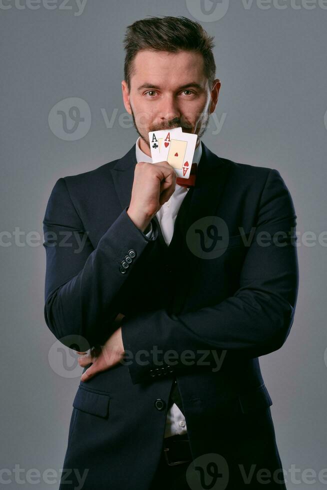 Man in black classic suit and red bow-tie showing two playing cards while posing against gray studio background. Gambling, poker, casino. Close-up. photo