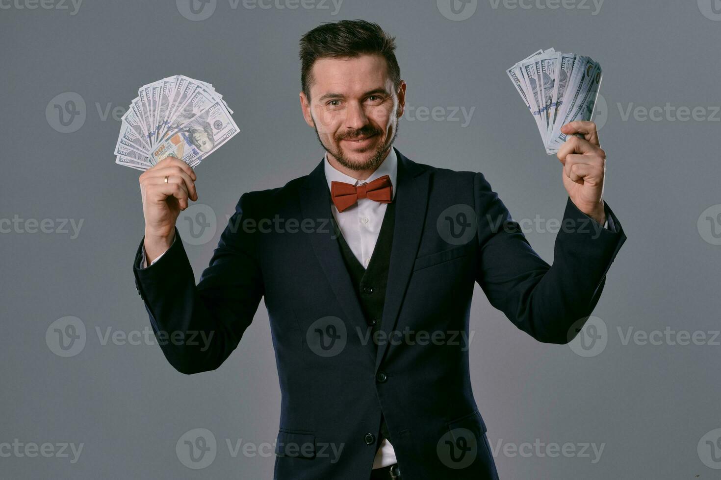 hombre en negro traje y rojo corbata de moño es demostración dos aficionados de cien dólar facturas, posando en gris estudio antecedentes. juego, póker, casino. de cerca. foto