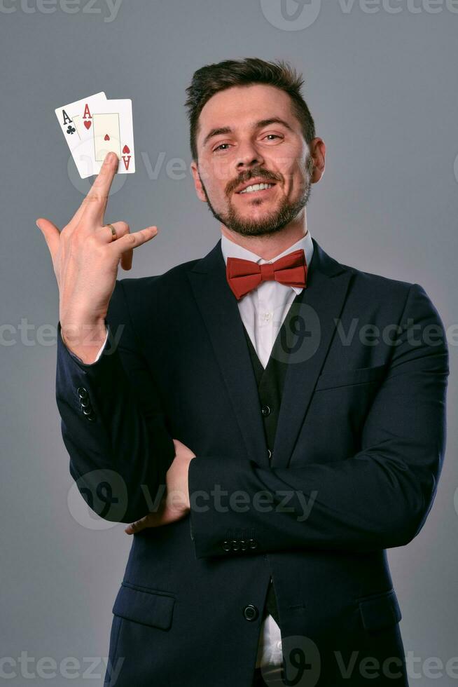 Man in black classic suit and red bow-tie showing two playing cards while posing against gray studio background. Gambling, poker, casino. Close-up. photo