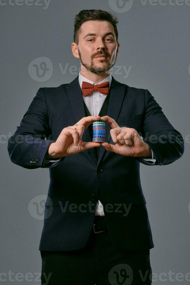 Man in black classic suit and red bow-tie showing some colored chips, posing against gray studio background. Gambling, poker, casino. Close-up. photo