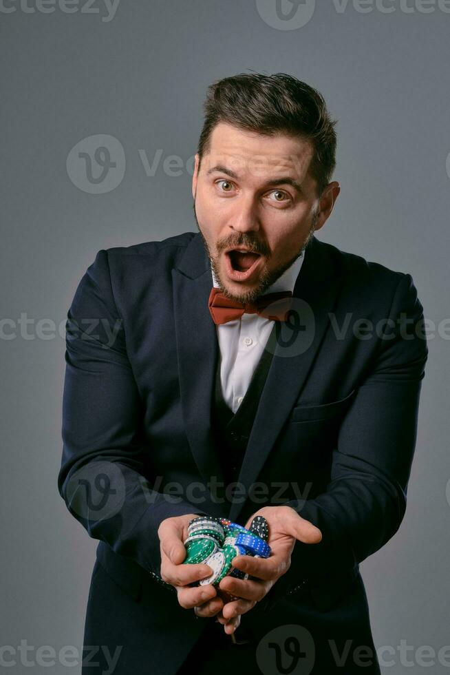 Man in black classic suit and red bow-tie showing some colored chips, posing against gray studio background. Gambling, poker, casino. Close-up. photo