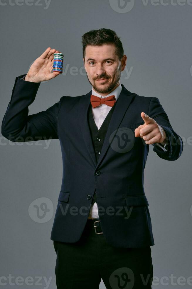 Man in black classic suit and red bow-tie showing some colored chips, posing against gray studio background. Gambling, poker, casino. Close-up. photo