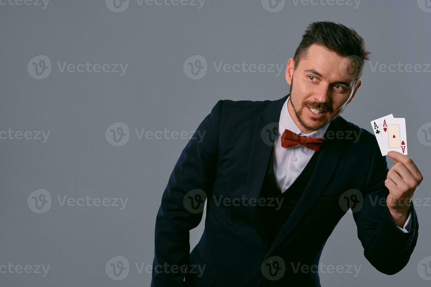 Man in black classic suit and red bow-tie showing two playing cards while posing against gray studio background. Gambling, poker, casino. Close-up. photo