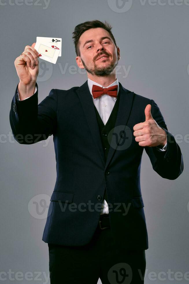 Man in black classic suit and red bow-tie showing two playing cards while posing against gray studio background. Gambling, poker, casino. Close-up. photo