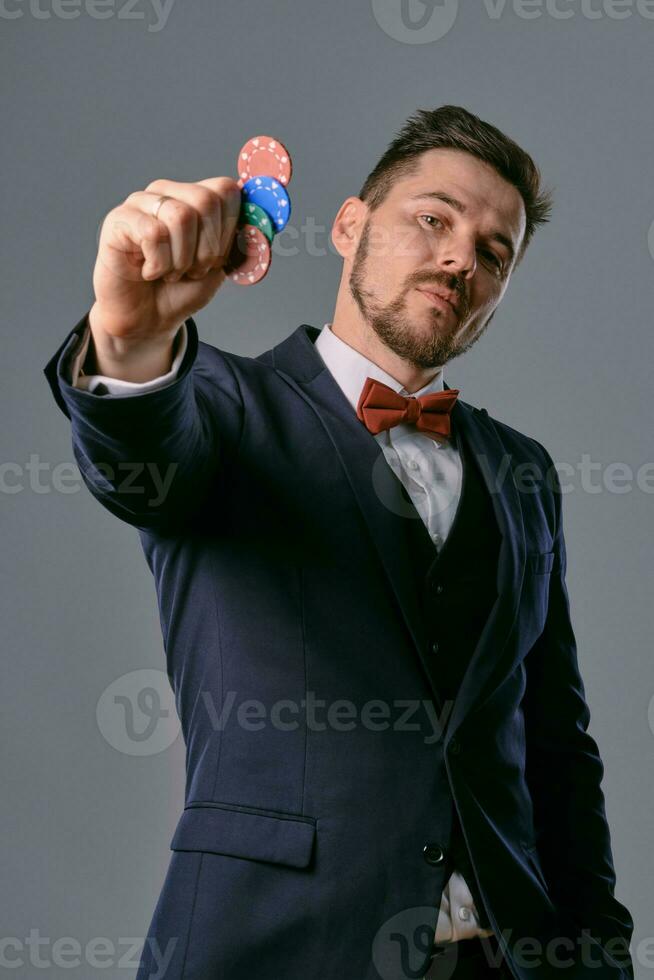 Man in black classic suit and red bow-tie showing some colored chips, posing against gray studio background. Gambling, poker, casino. Close-up. photo