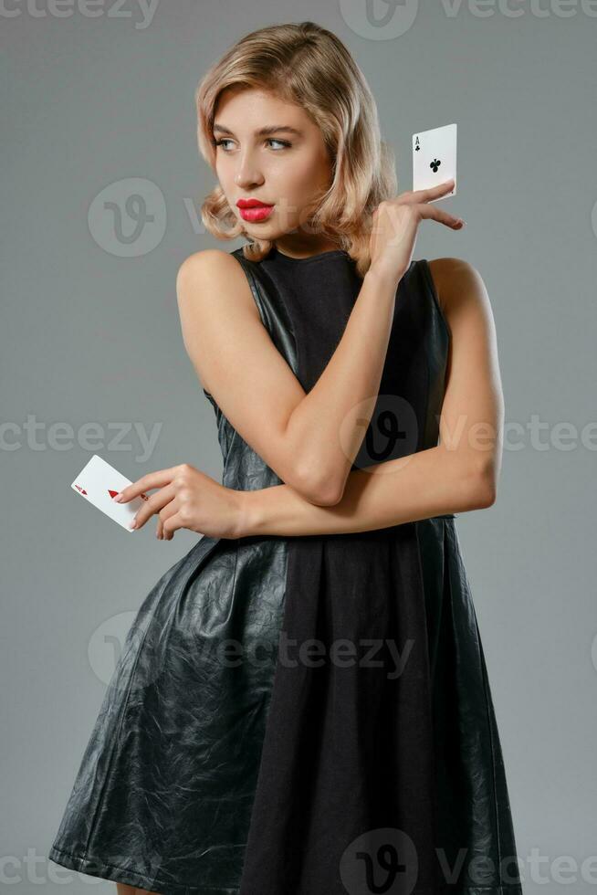 Blonde girl in black stylish dress showing two playing cards, posing against gray background. Gambling entertainment, poker, casino. Close-up. photo