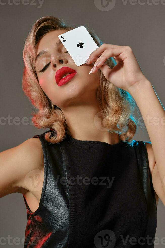 Blonde girl in black leather dress showing ace of clubs, posing against gray background. Gambling entertainment, poker, casino. Close-up. photo