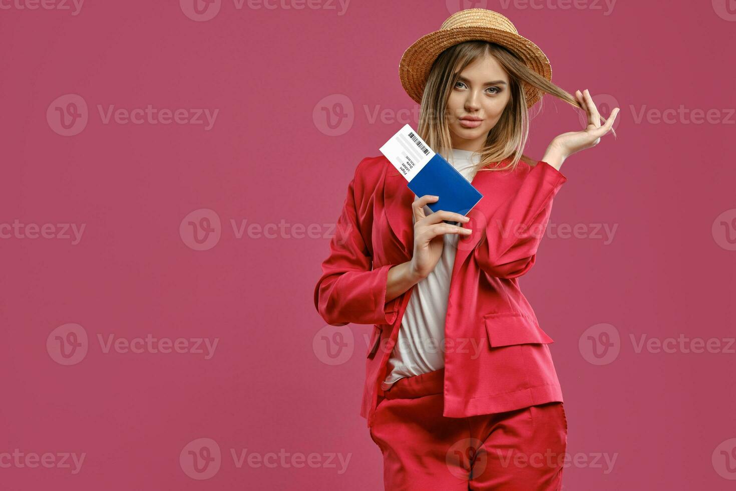 Blonde woman in straw hat, white blouse and red pantsuit. She is holding passport and ticket while posing against pink studio background. Close-up photo