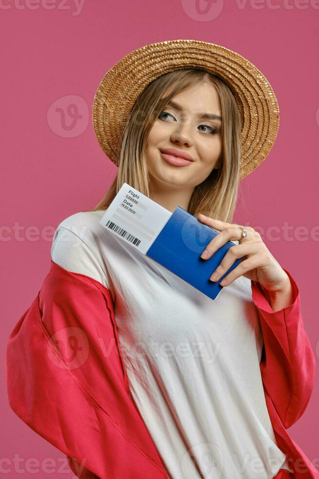 Blonde woman in straw hat, white blouse and red pantsuit. She smiling, holding passport and ticket while posing on pink studio background. Close-up photo