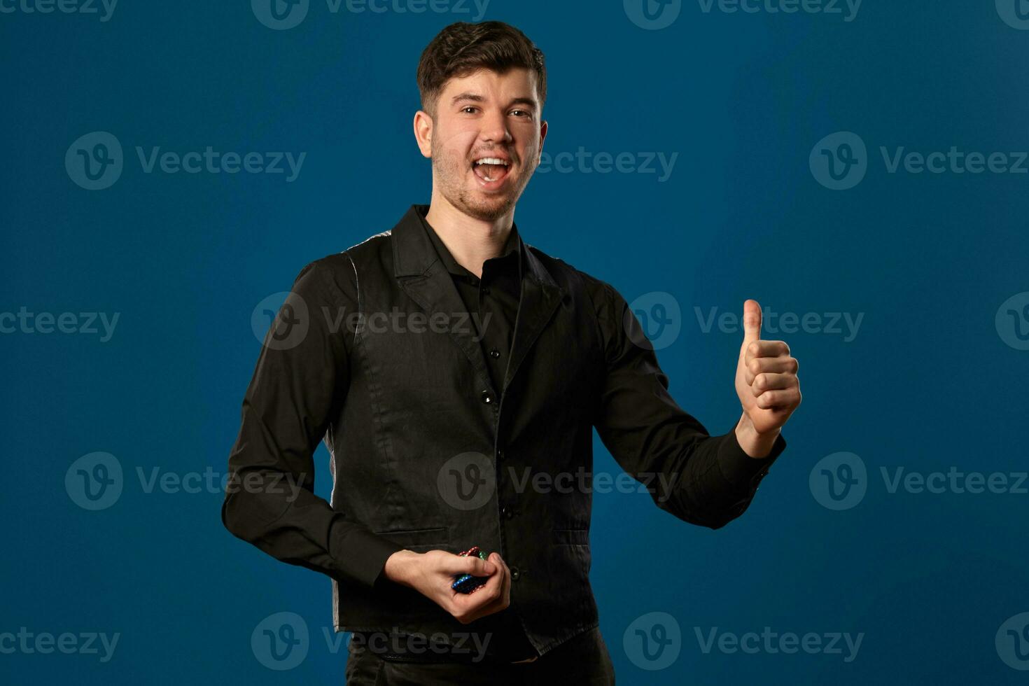 Newbie in poker, in black vest and shirt. Holding some colored chips. Posing against blue background. Gambling, casino. Close-up. photo