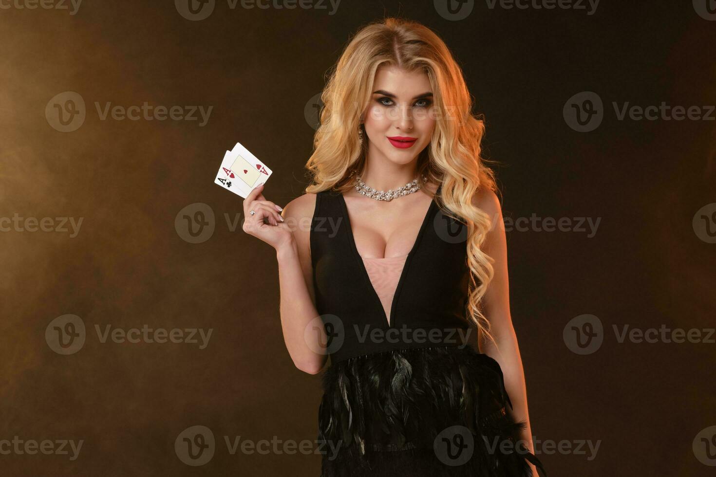 Blonde lady in black dress with feathers and necklace. She is smiling, showing two aces, posing on black smoky background. Poker, casino. Close-up photo