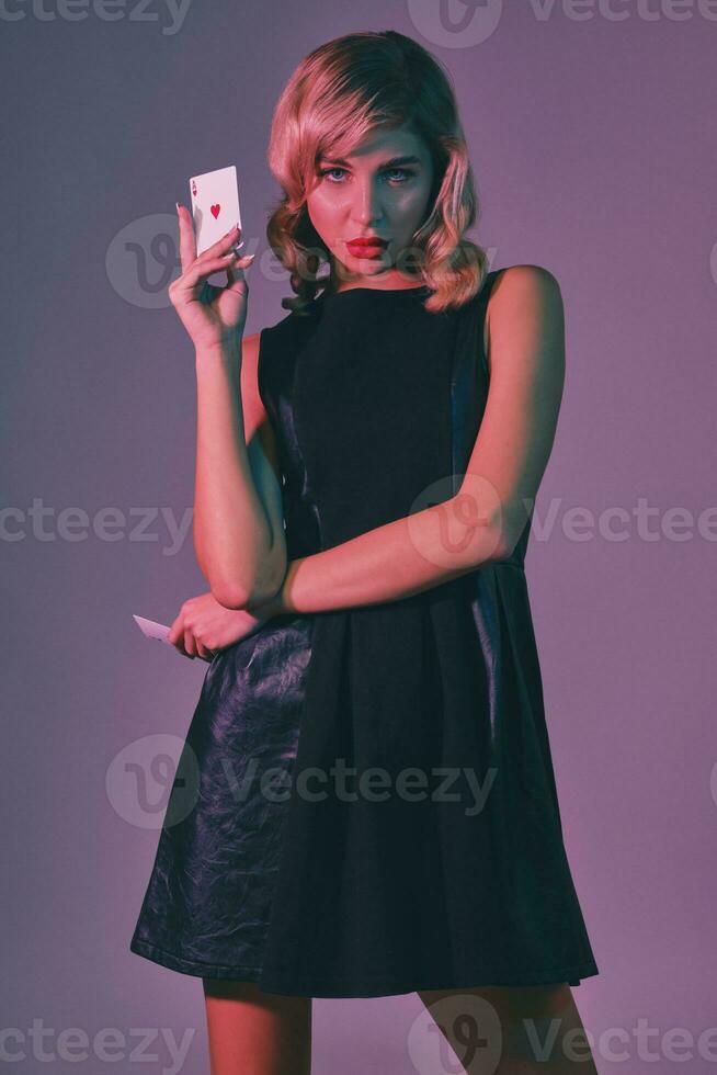 Blonde girl in black stylish dress showing two playing cards, posing against colorful background. Gambling entertainment, poker, casino. Close-up. photo