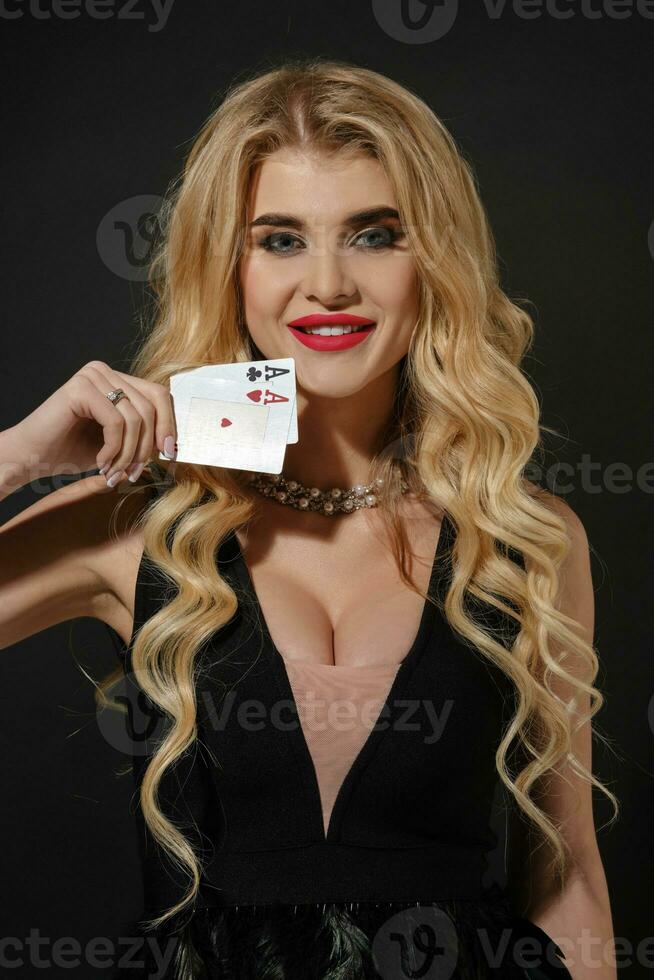Blonde female in black stylish dress and necklace. She smiling, showing two playing cards, posing on black background. Poker, casino. Close-up photo