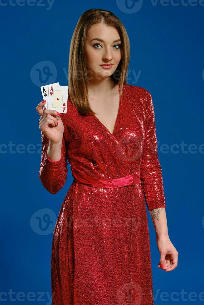 Blonde woman with make-up, tattooed hand, in red sequin dress is showing two aces, posing on blue background. Gambling, poker, casino. Close-up. photo