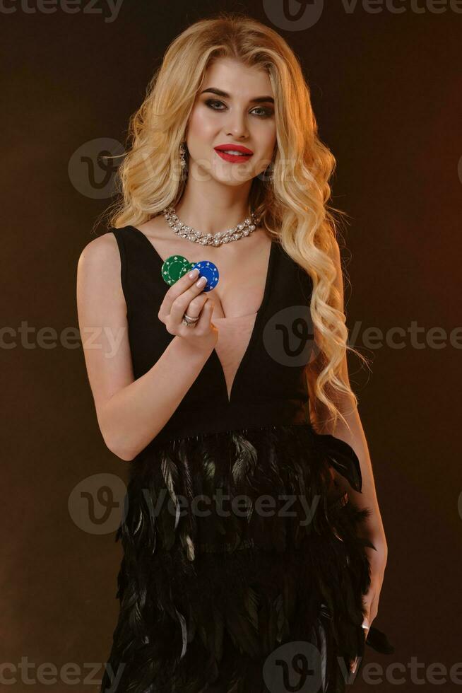Blonde lady in black dress with feathers and necklace. She smiling, showing green and blue chips, posing on brown background. Poker, casino. Close-up photo