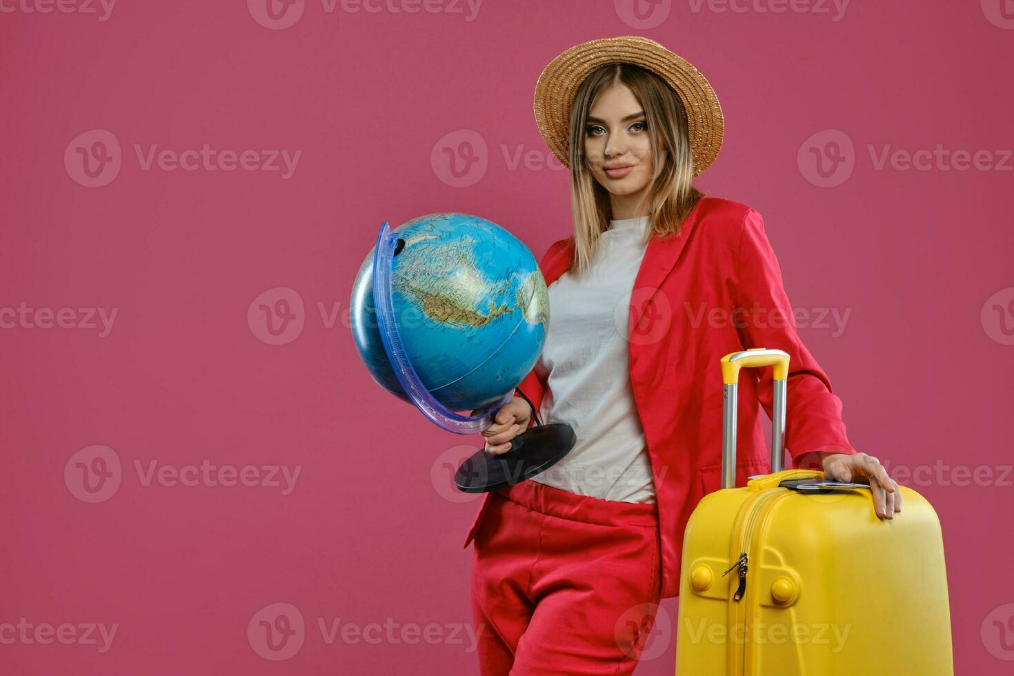 Lady in hat, white blouse, red pantsuit. Smiling, holding globe. Leaning on yellow suitcase, passport and ticket on it, posing on pink background photo