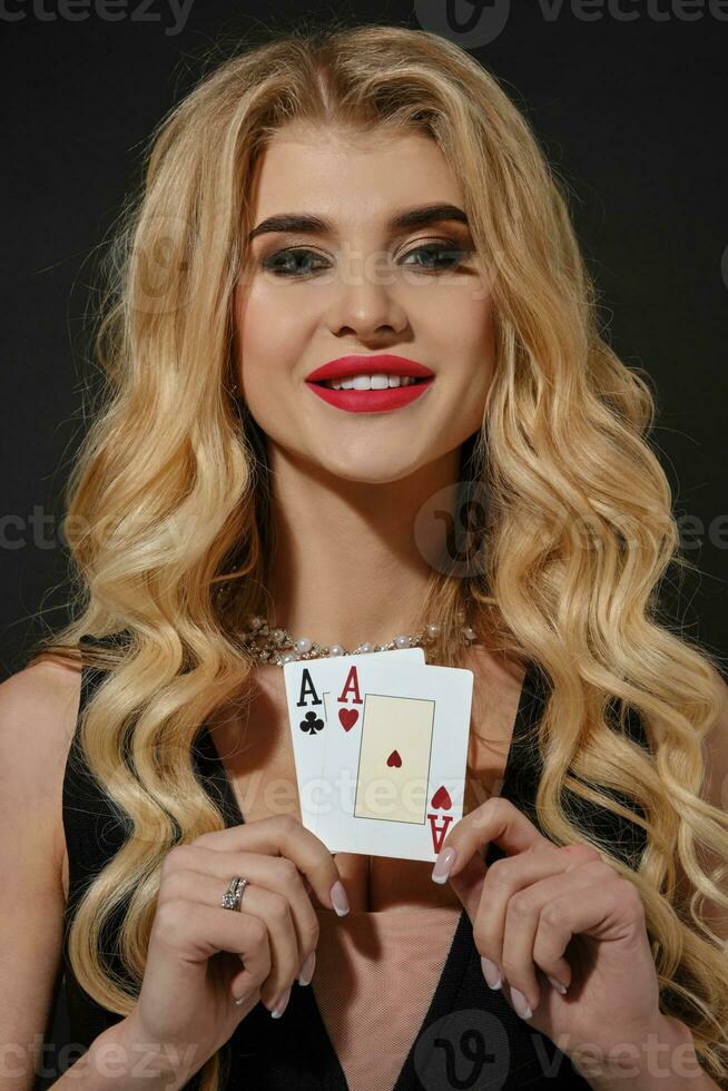 Blonde girl in black stylish dress and necklace. She smiling, showing two aces, posing on black studio background. Poker, casino. Close-up photo