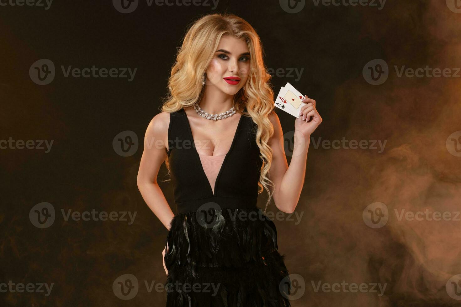 Blonde lady in black dress with feathers and necklace. She is smiling, showing two aces, posing on black smoky background. Poker, casino. Close-up photo