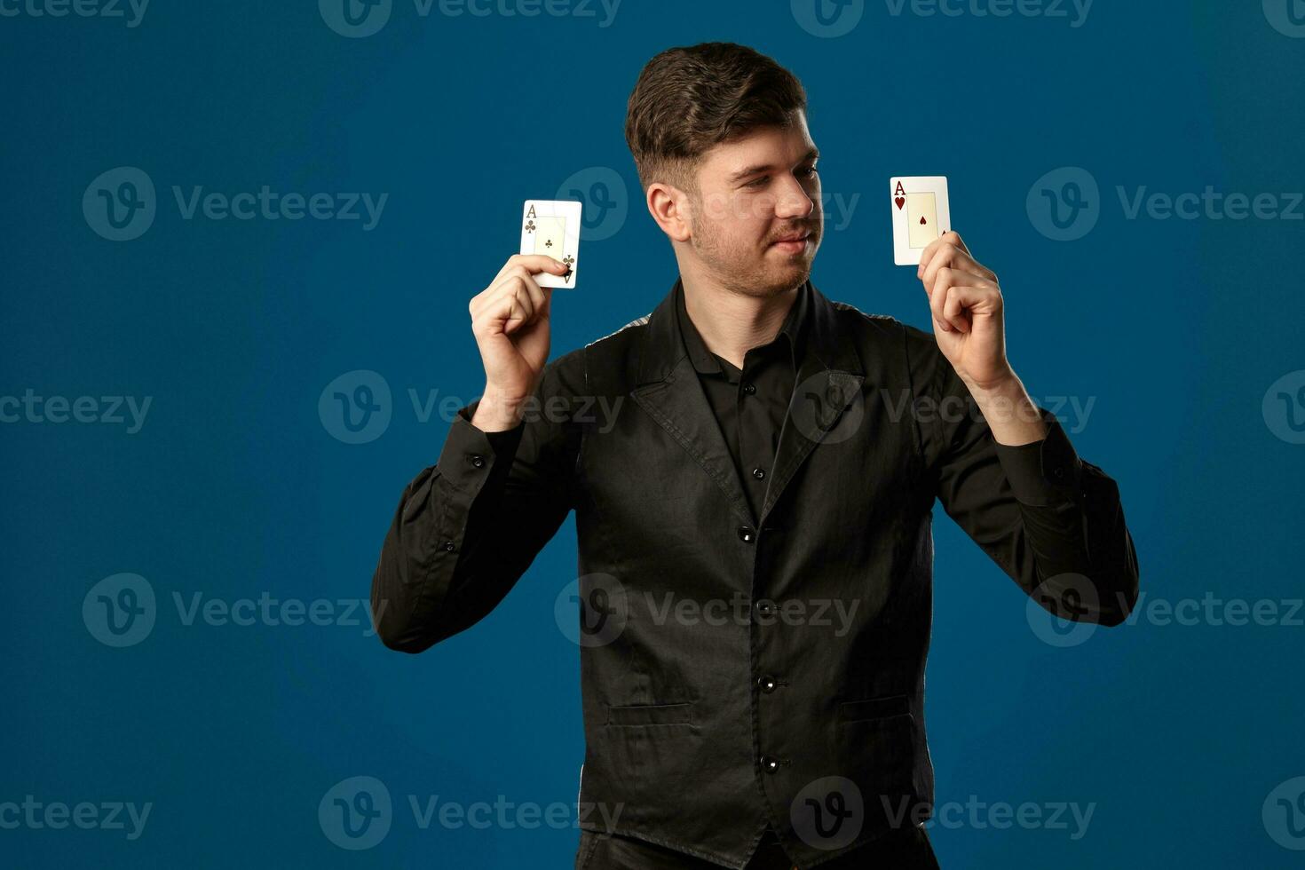 Noob in poker, in black vest and shirt. Holding two playing cards while posing against blue studio background. Gambling, casino. Close-up. photo