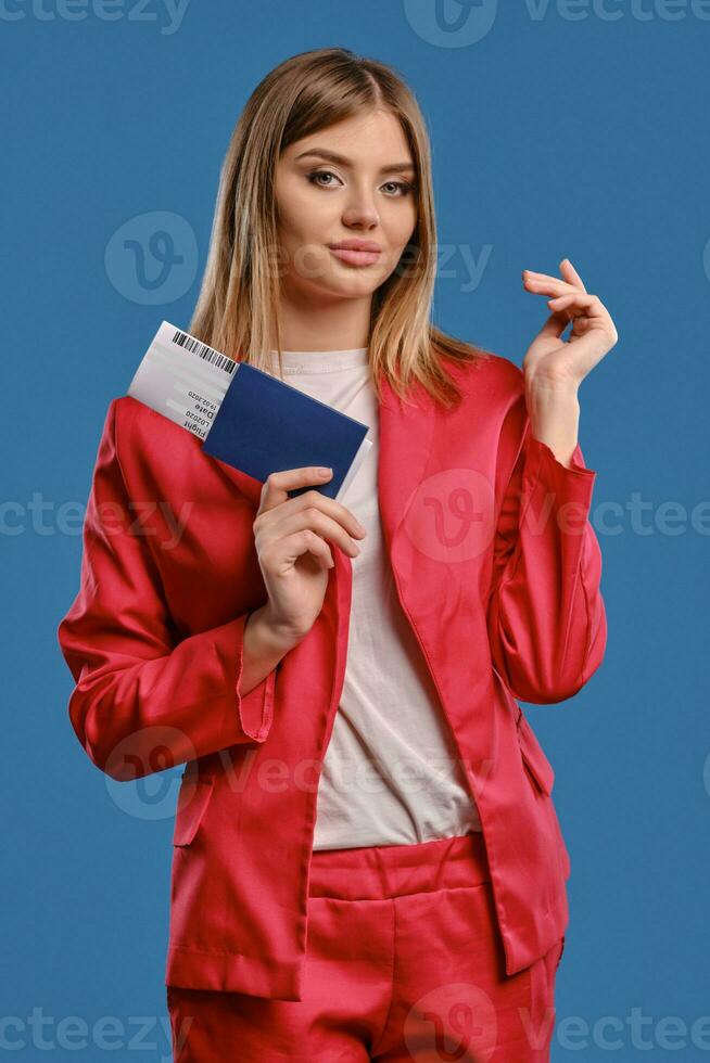 Blonde female in white blouse and red pantsuit. She smiling, holding passport and ticket while posing on blue studio background. Close-up photo