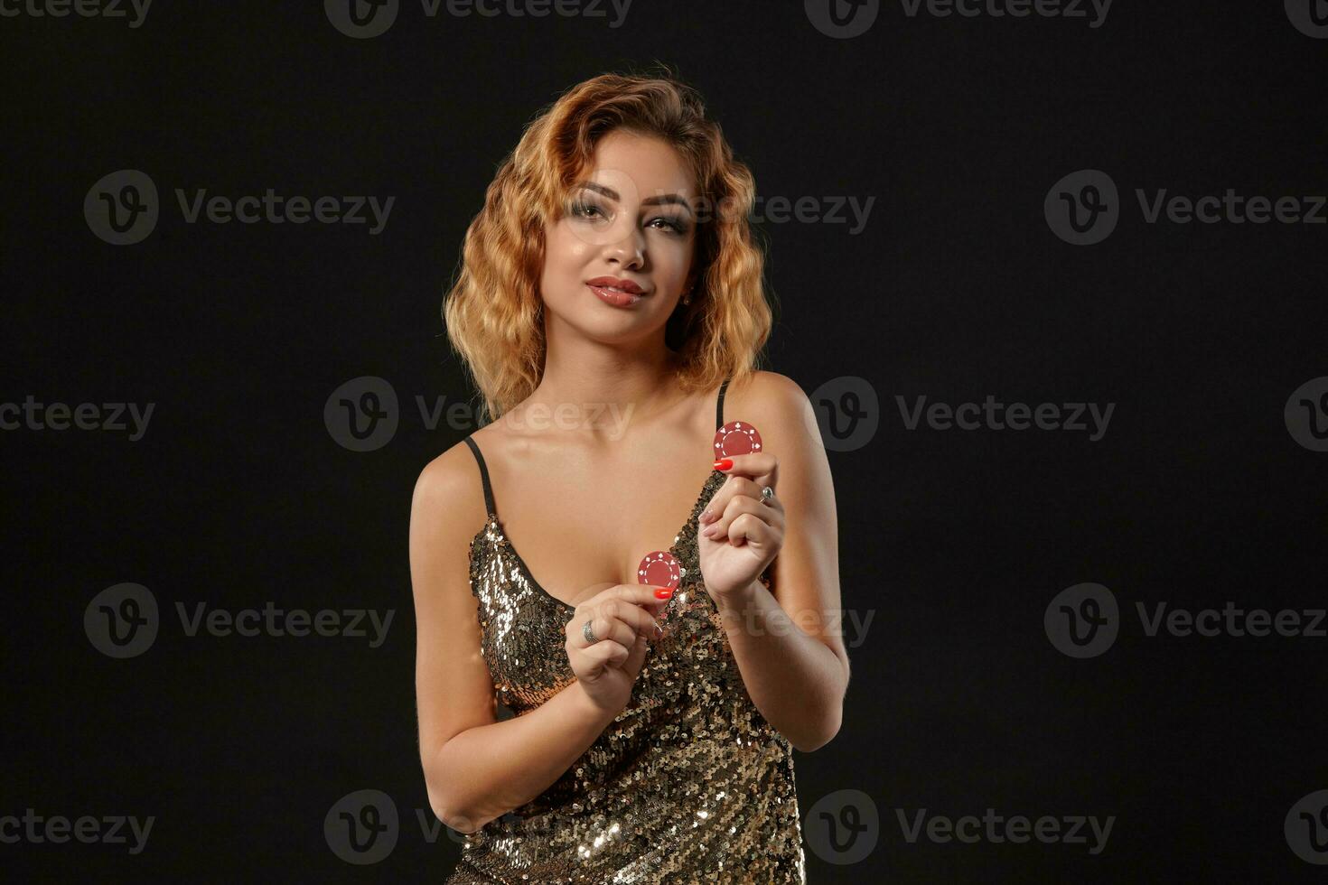 Ginger girl wearing shiny dress is posing holding two red chips in her hands standing against a black studio background. Casino, poker. Close-up. photo