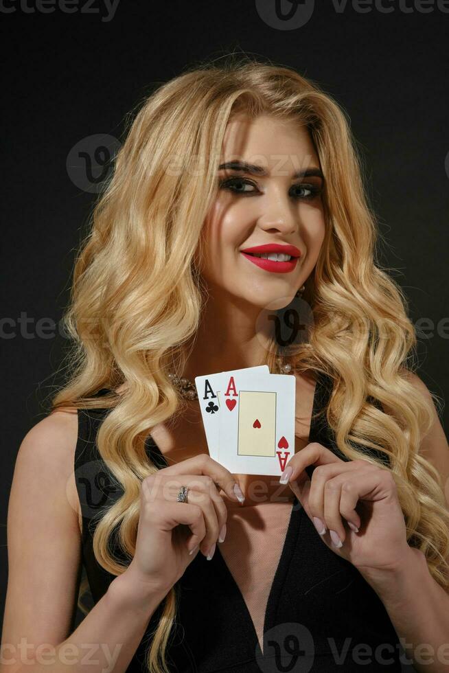 Blonde model in black stylish dress and necklace. She smiling, showing two aces, posing on black background. Poker, casino. Close-up photo