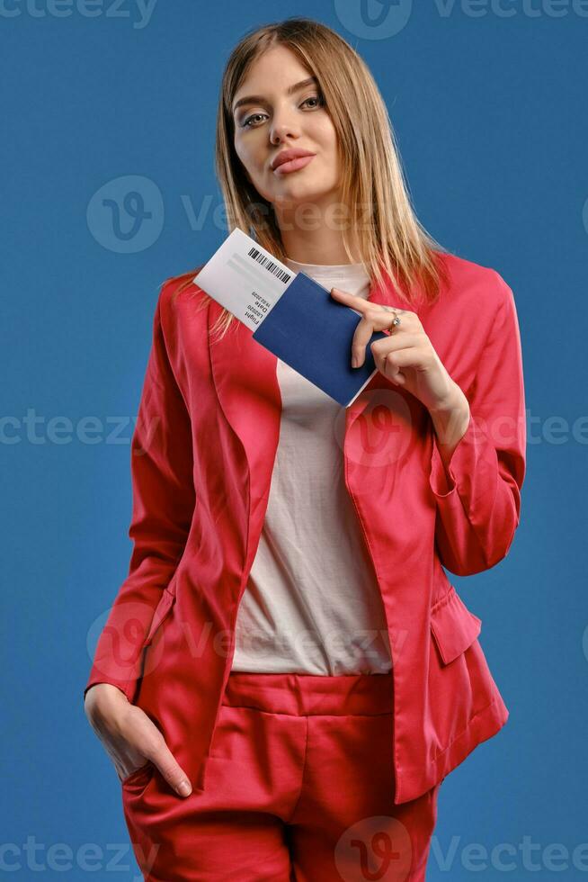 Blonde lady in white blouse and red pantsuit. She smiling, holding passport and ticket while posing on blue studio background. Close-up photo