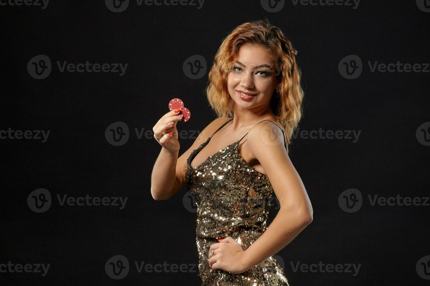 Ginger girl wearing shiny dress is posing holding two red chips in her hands standing against a black studio background. Casino, poker. Close-up. photo