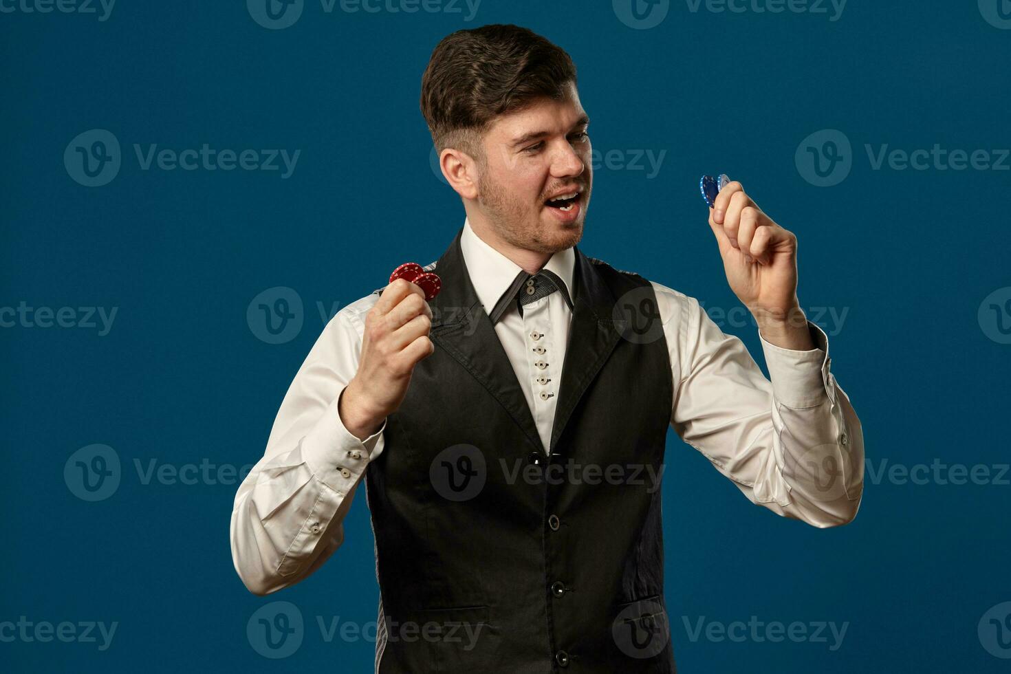 Newbie in poker, in black vest and white shirt. Holding some colored chips. Posing against blue background. Gambling, casino. Close-up. photo