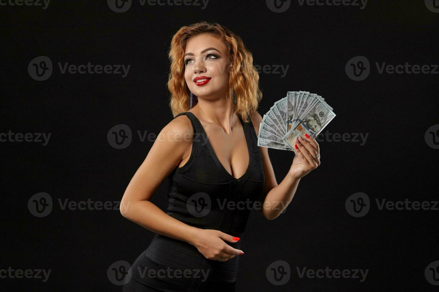 Ginger girl in dark dress posing holding fan of hundred-dollar bills in her hands standing against a black studio background. Casino, poker. Close-up. photo