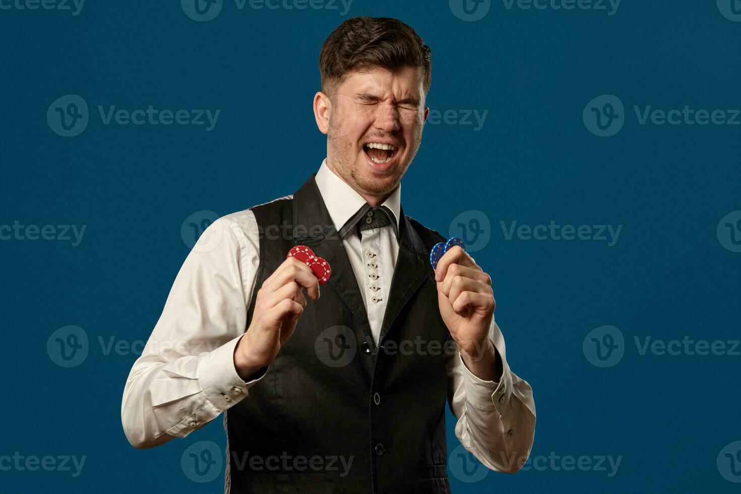 Newbie in poker, in black vest and white shirt. Holding some colored chips. Posing against blue background. Gambling, casino. Close-up. photo