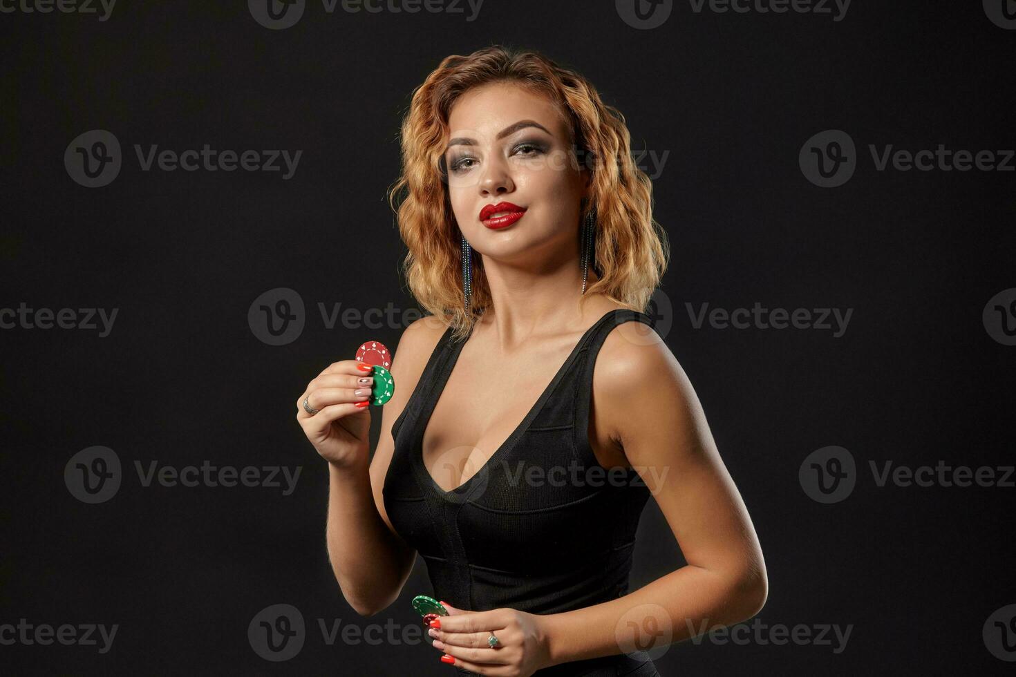 Ginger girl wearing dark dress is posing holding red and green chips in her hands standing against black studio background. Casino, poker. Close-up. photo