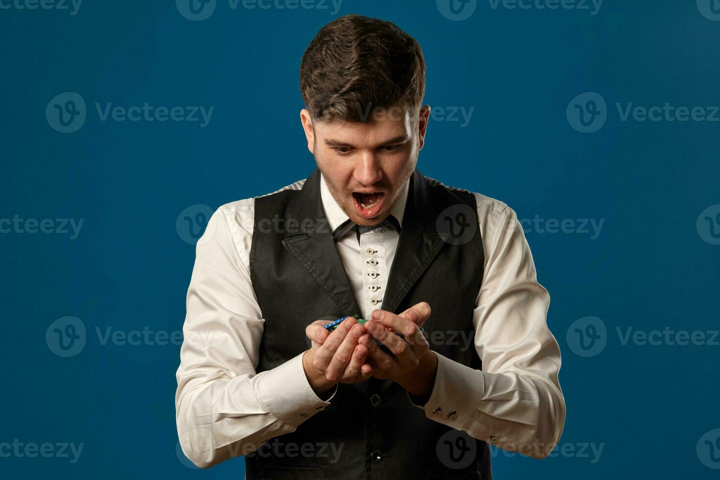 Newbie in poker, in black vest and white shirt. Holding some colored chips. Posing against blue background. Gambling, casino. Close-up. photo