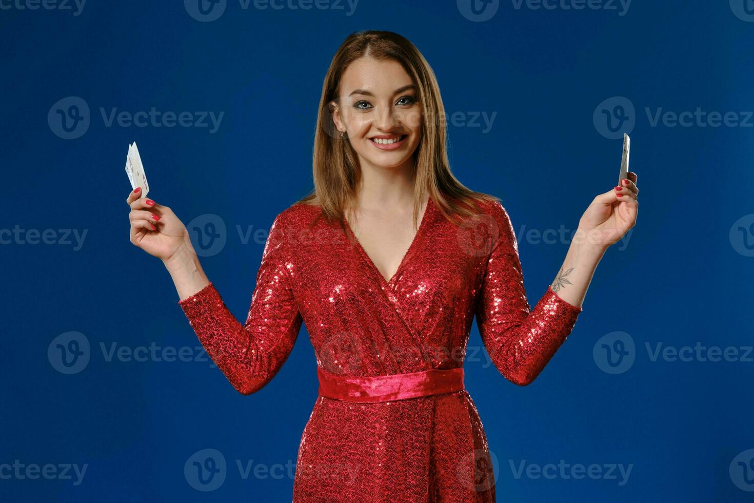 Blonde model with make-up, tattooed hand, in red sequin dress is smiling, showing four aces, posing on blue background. Poker, casino. Close-up. photo