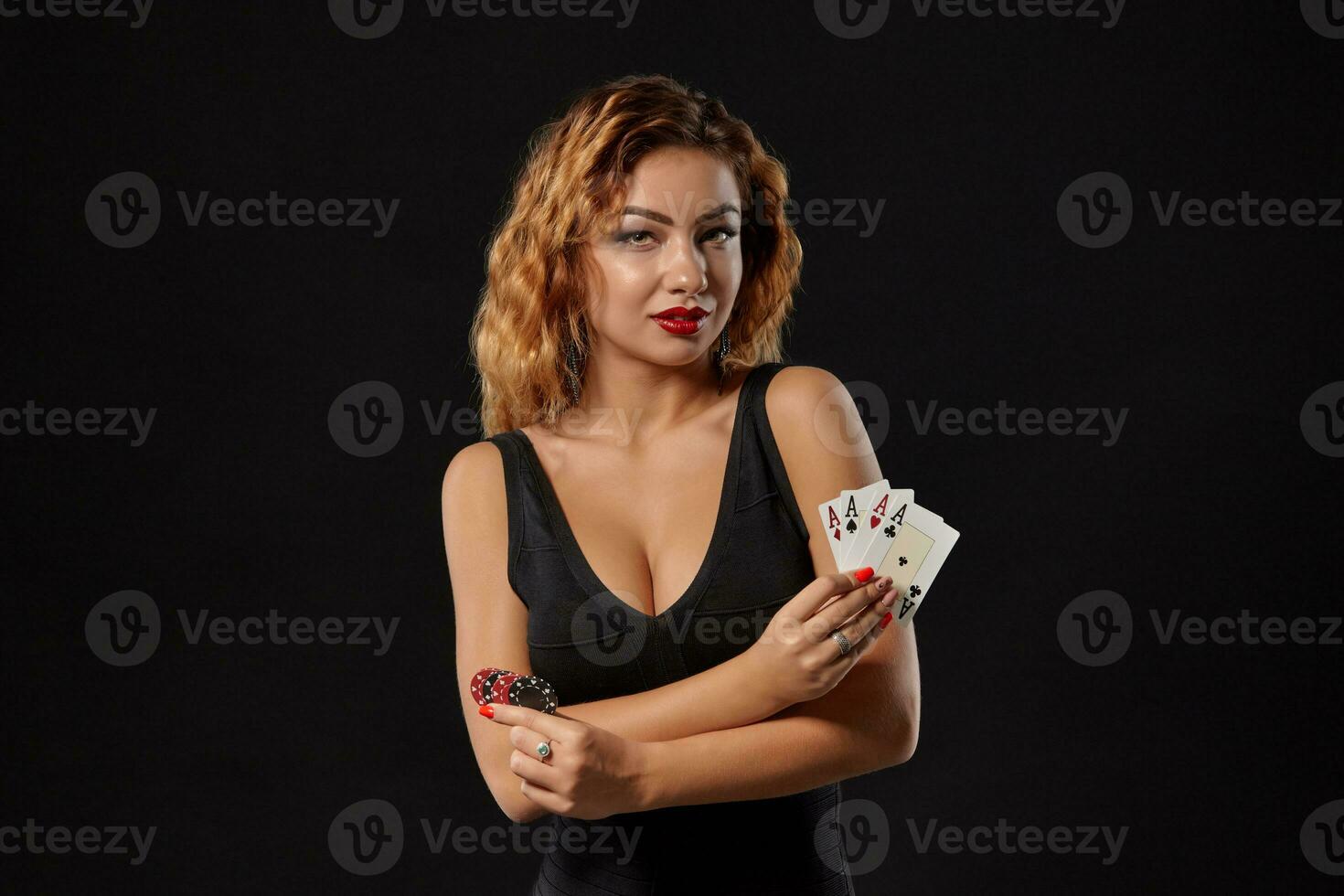 Ginger girl in a dark dress posing holding playing cards and chips in her hands standing against a black studio background. Casino, poker. Close-up. photo