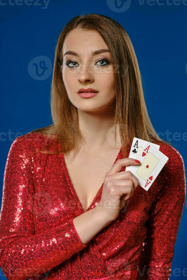 Blonde female with make-up, in red sequin dress is showing two aces, posing on blue background. Gambling, poker, casino. Close-up. photo