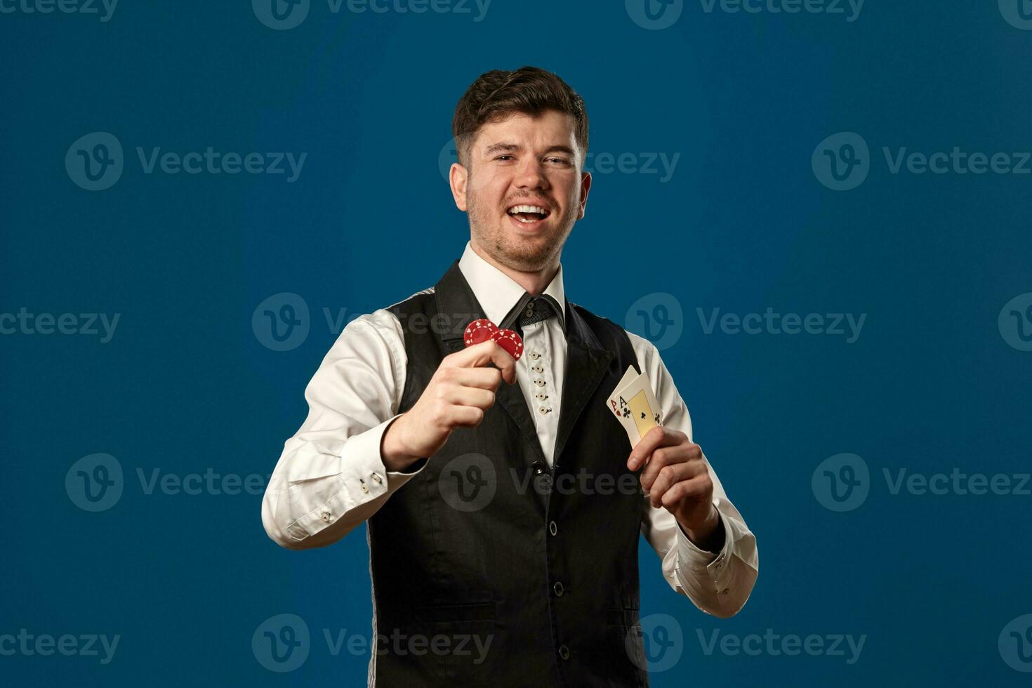 Noob in poker, in black vest and white shirt. Holding two red chips and aces. Posing against blue background. Gambling, casino. Close-up. photo