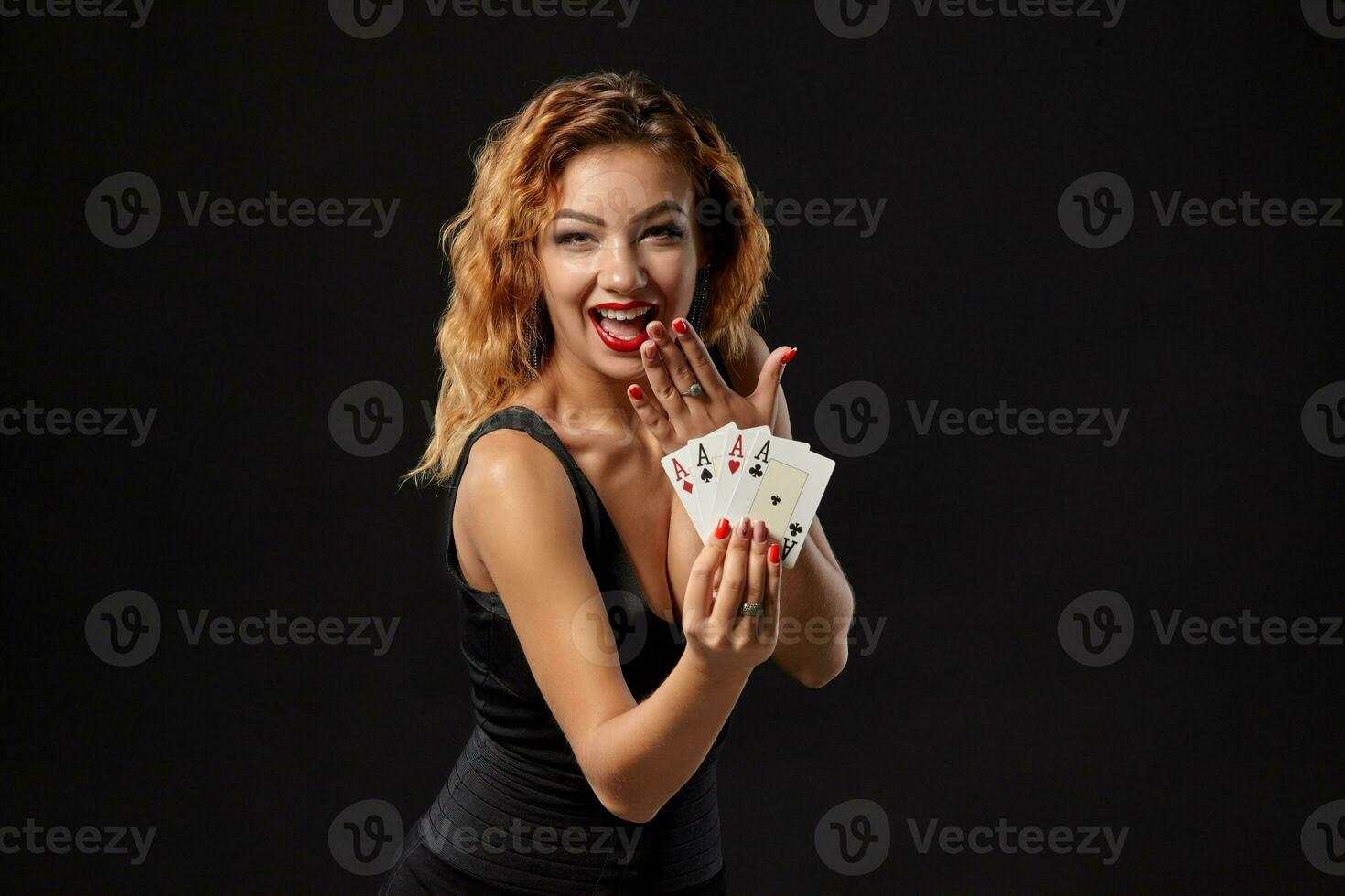 Ginger girl wearing a dark dress is posing holding four aces in her hands standing against a black studio background. Casino, poker. Close-up. photo