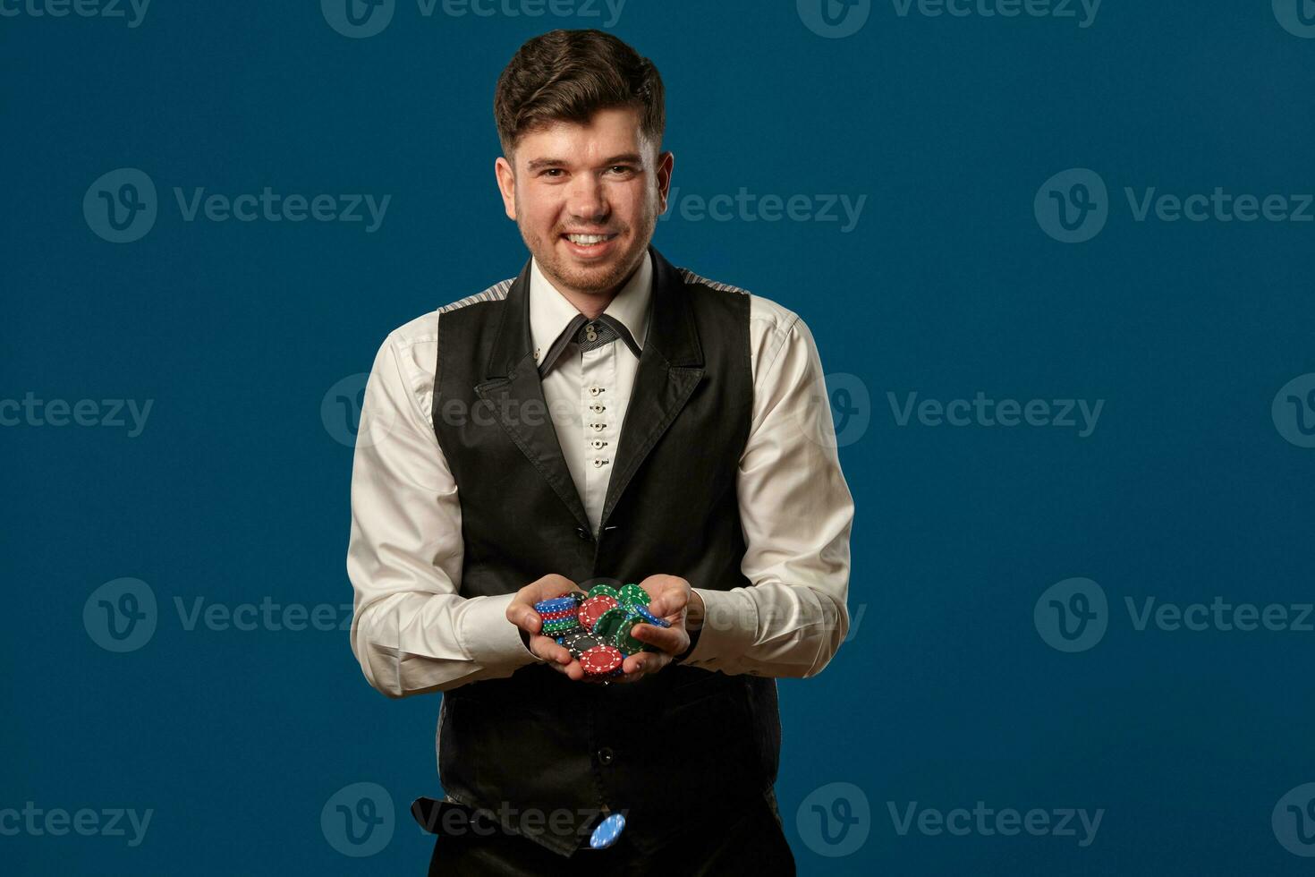 Noob in poker, in black vest and trousers, white shirt. Holding some colored chips. Posing against blue background. Gambling, casino. Close-up. photo