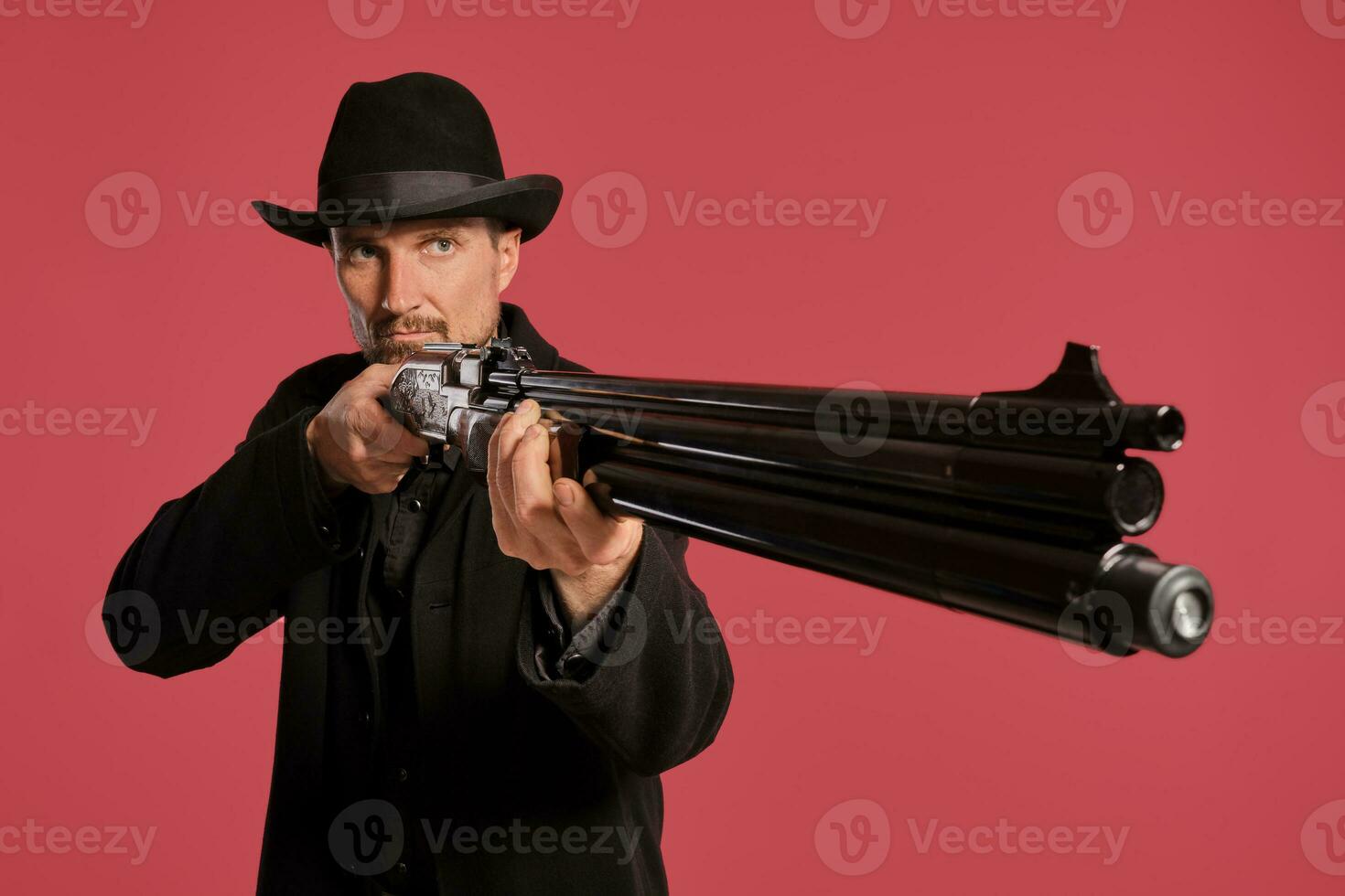 Middle-aged man with beard, mustache, in black jacket and hat, holding a gun while posing against a red background. Sincere emotions concept. photo