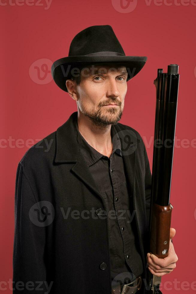 de edad mediana hombre con barba, Bigote, en negro chaqueta y sombrero, participación un pistola mientras posando en contra un rojo antecedentes. sincero emociones concepto. foto